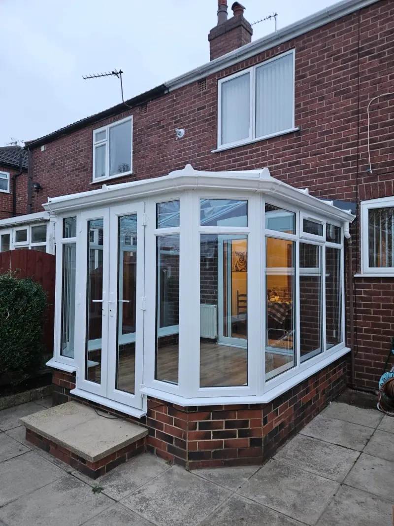 A white-framed lean-to conservatory on the back of a red brick house, with a splayed bay front, a pair of glazed double doors on the left, opening toplights above the fixed panes, and a low brick dwarf wall under the glass. Inside are wood-effect flooring, a white radiator and a wooden chair. Paving slabs and a concrete step run up to the doors.