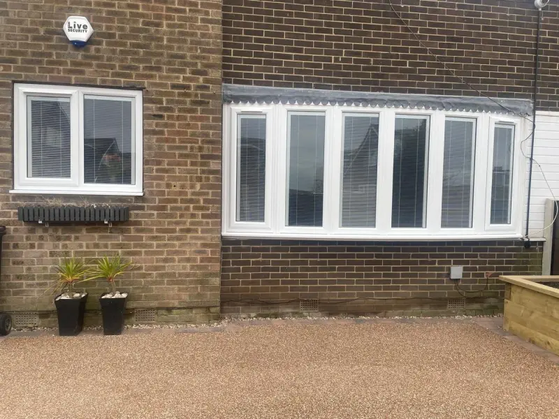A six-light white uPVC bow window with integral blinds set in dark brick, with a separate two-pane casement window on the adjoining wall, above a resin-bound driveway. An alarm box lettered Live SECURITY is fixed high on the brick, and a dark column radiator is mounted on the outside wall below the casement.