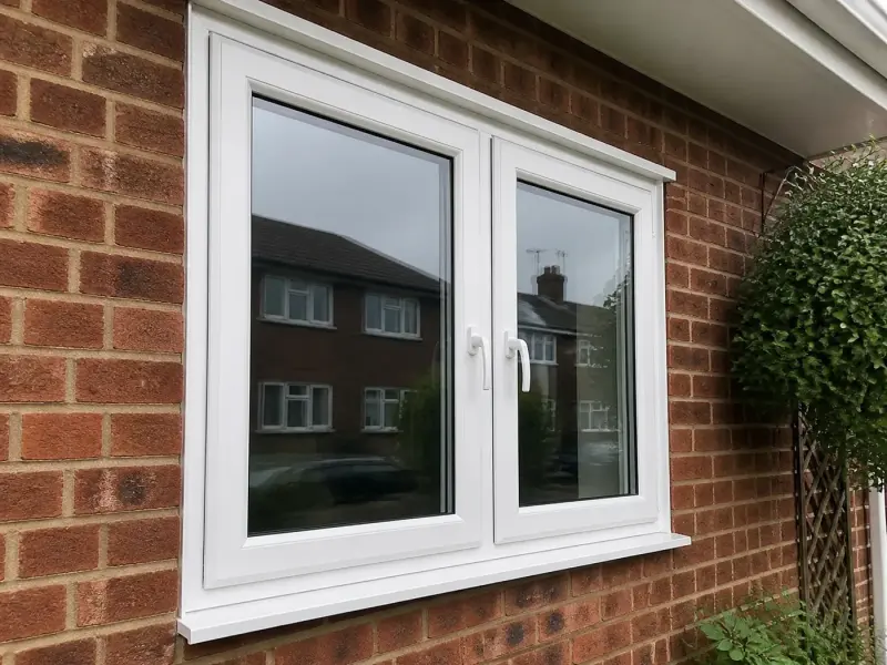 Illustrative image of a white uPVC two-casement double-glazed window set in red brick, with neighbouring houses reflected in the glass and a clipped shrub beside it.