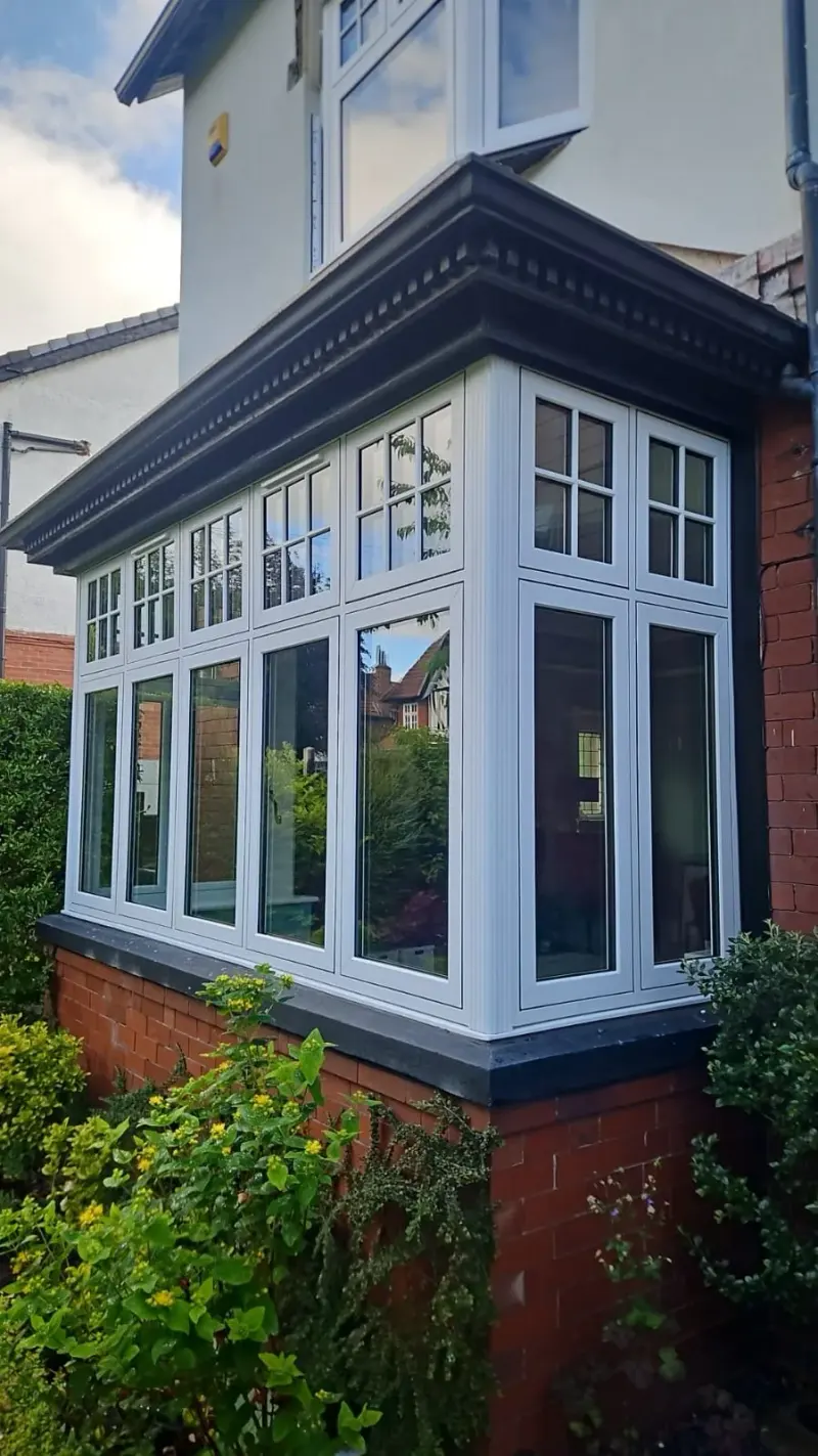 A large square bay window run in white uPVC with Georgian-bar toplights, wrapping two elevations above a red-brick dwarf wall, beneath a dentil-moulded cornice.